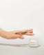 A person's hand holding a small jar of 'reBRAND' brand moisturizer, with the jar placed on a light surface.